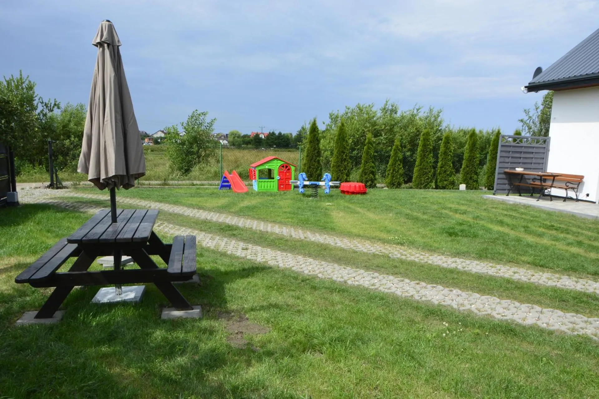 Plac zabaw w ogrodzie — zjeżdżalnia, domek zabawowy, bujaki i piaskownica, obok stół piknikowy z parasolem