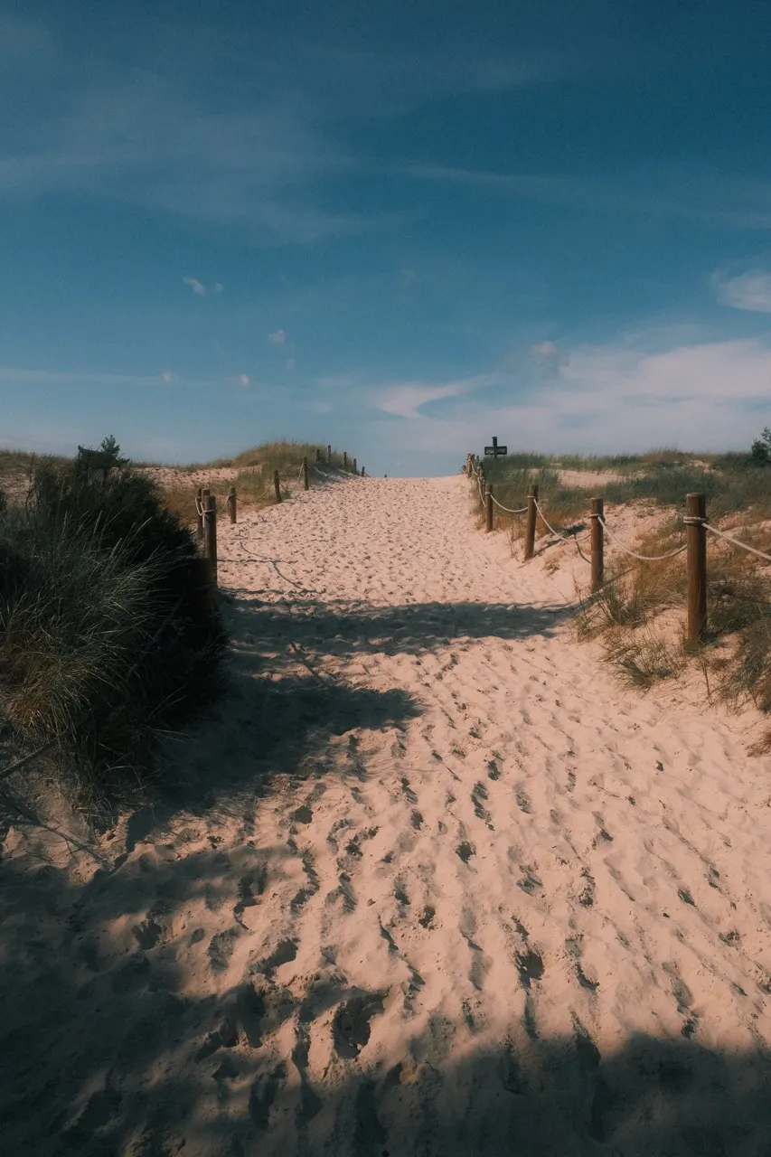 Piaszczyste zejście na plażę w Łebie między porośniętymi trawą wydmami, wzdłuż sznurowej barierki
